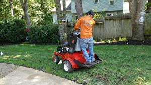 A man in an orange shirt is standing on the back of a lawn mower.